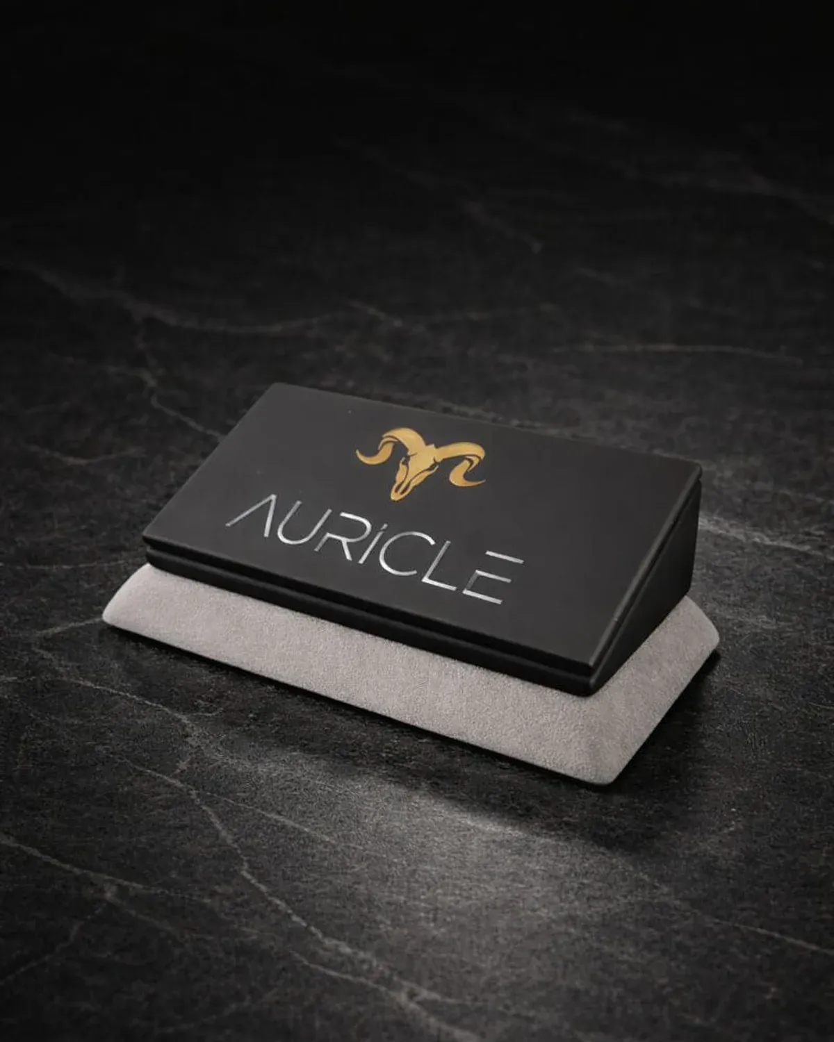 AURICLE jewellery branded nameplate stand. The stand has a light gray suede base with a black leather top. Sat on a dark worktop with moody lighting.