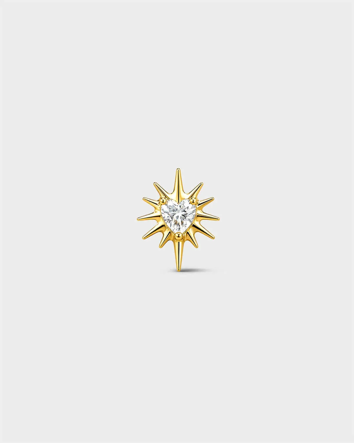 Gold titanium threadless end earring with a heart-cut clear stone set within a sculpted starburst design. On a light grey background.